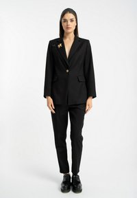 Czarny dopasowany garnitur składający się z dwurzędowego żakietu z złotym guzikiem i dopasowanych spodni wąskich, połączony z masywnymi czarnymi butami.