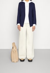 Blazer azul marino con una sudadera blanca con un logo, pantalones de pierna ancha color crema y un bolso de piel beige. Unos zapatos negros de suela gruesa completan el look.