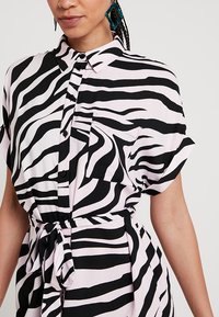 Czarna i biała sukienka koszulowa w paski zebra, wykonana z lekkiego materiału. Posiada kołnierzyk, krótkie rękawy, kieszeń z przodu oraz sznurek w talii.