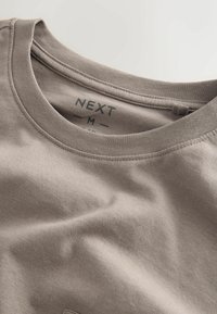 Maglietta beige a maniche corte con scollo rotondo. Il materiale sembra morbido con una texture liscia. L'etichetta mostra "NEXT" e la taglia "M".