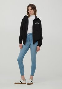 Felpa nera con zip e tasche frontali, con la scritta "New York Athletic", abbinata a jeans skinny azzurri chiari e sneakers bianche con accenti neri.