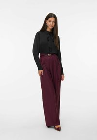 Zwarte blouse met knoopsluiting en strikdetail, gecombineerd met hooggeremde, wijd-uitlopende bordeauxrode broek, voorzien van een glanzende riem en spitse schoenen.