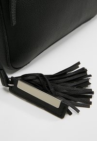 Black textured leather bag corner with attached black leather tassel featuring a rectangular silver metal accent on white background.