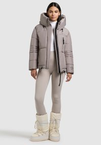 Lichtgrijze pufferjack met een grote capuchon, zwarte rits, voorzakken en horizontale stiksels. Gecombineerd met beige leggings en witte laarzen.