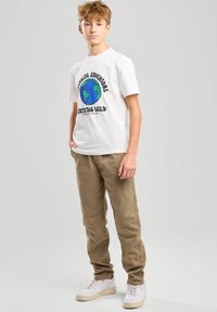 Camiseta de algodón blanca con un gráfico de globo azul y verde y texto en negro, combinada con pantalones beige y zapatillas blancas.