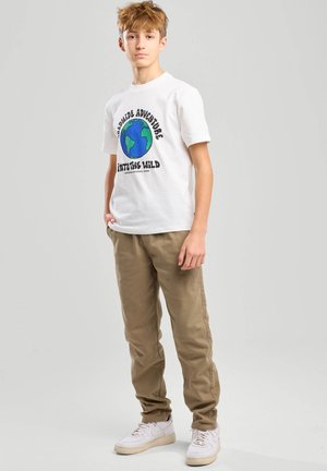 Ragazzo adolescente in piedi con una t-shirt bianca con grafica di un globo e testo, pantaloni beige e scarpe da ginnastica bianche, su uno sfondo semplice.