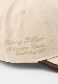 Beige canvas baseball cap with brown leather trim and embroidered text reading "Tommy Hilfiger 5th American Classic Established 1985."