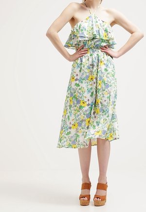 Frau trägt ein neckholder Blumenkleid mit Rüschendetails und braunen Plateau-Sandalen, steht mit den Händen in der Hüfte.