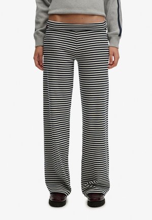 Vrouw draagt losse zwart-witte broek met horizontale strepen en donkerbruine schoenen, staande met ontspannen handen langs het lichaam.