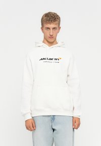 Ung mand iført en hvid McLaren Formel 1 Team hoodie og lyseblå jeans, stående imod en ensfarvet hvid baggrund.