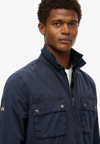 Navy blue jacket with a high collar, front zipper, and two buttoned chest pockets. Features a textured fabric and subtle logo detail.