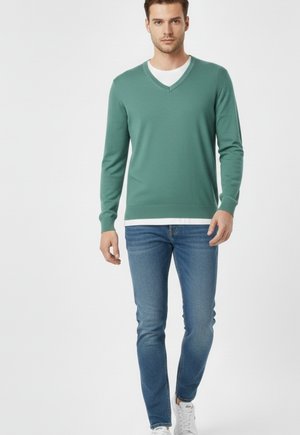 Suéter verde de punto con cuello en V y mangas largas, combinado con jeans ajustados azules. El modelo lleva zapatillas blancas y una camiseta interior blanca.