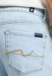 Jasnoniebieskie dżinsy mają tylnią kieszeń z żółtym szwem w falisty wzór. Widać również granatową metkę z białym napisem.