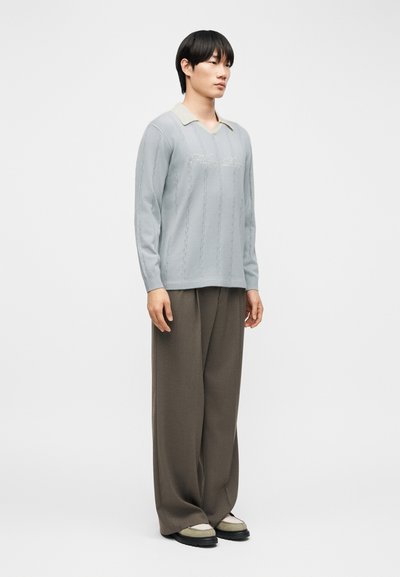 Νέος άνδρας που στέκεται φορώντας ανοιχτό μπλε μακρυμάνικη μπλούζα πόλο και φαρδιά καφέ παντελόνια με μπεζ και μαύρα παπούτσια, κοιτάζοντας προς τα δεξιά.