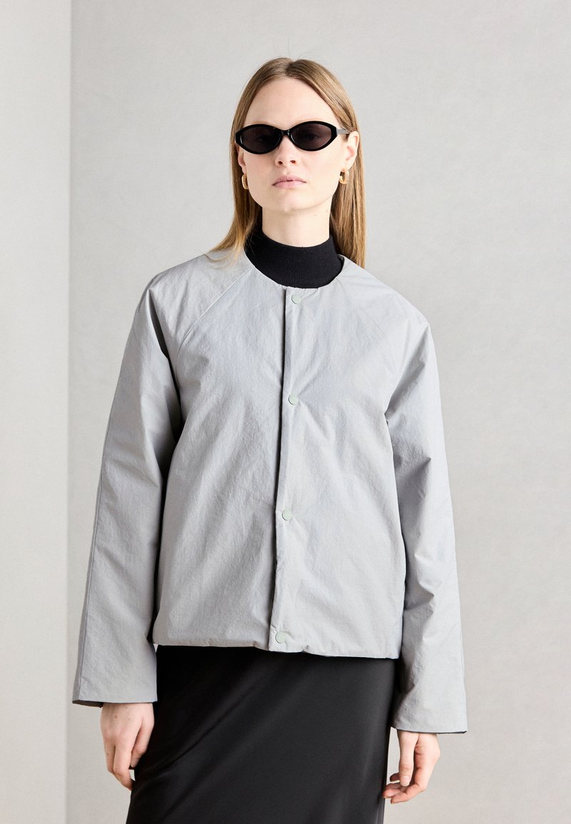 Lys grå jakke med rund krage, trykknaplukking og lange ermer. Brukt over en svart turtleneck, kombinert med stilige svarte solbriller.