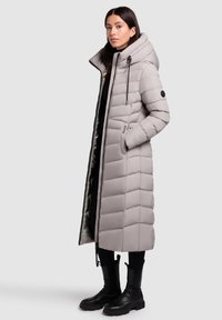 Manteau d'hiver gris en forme de puffer avec une silhouette longue, motif matelassé, capuche amovible, fermeture éclair noire et poches latérales, porté sur des vêtements et des bottes noires.