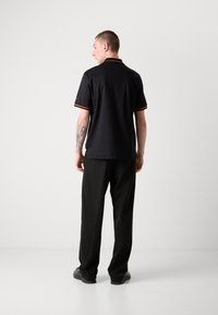 Polo noir à manches courtes, avec des bandes d'accent rouge sur le col et les manches, associé à un pantalon noir et des chaussures grises.