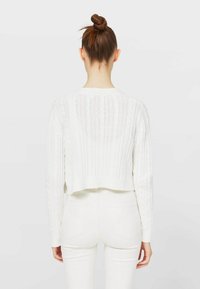 Bílý krátký svetr s vertikálním žebrovaným vzorem, dlouhými rukávy a kulatým výstřihem, doplněný bílými přiléhavými kalhotami.