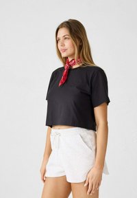 Camiseta corta negra con mangas cortas, combinada con pantalones cortos gris claro. La camiseta presenta un logo sutil y una bufanda roja alrededor del cuello.