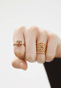 Dos anillos en una mano: una banda de oro con una piedra de corte esmeralda colorida y un anillo de oro con textura de trenzado. Ambos anillos presentan diseños intrincados.