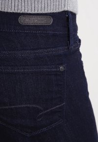 Jeans de mezclilla azul oscuro con un acabado texturizado, que cuentan con un bolsillo trasero, un parche de cuero en la cintura y sutiles detalles de costura.