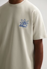 Camiseta de algodón color crema con mangas cortas. Presenta un diseño floral bordado en azul y un logo en el área del pecho izquierdo. Textura suave.