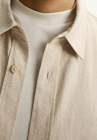 Beżowa koszula zapinana na guziki, wykonana z teksturowanej tkaniny, narzucona na jasnoszary sweter z okrągłym dekoltem. Posiada pojedyncze zapięcie na guzik i kołnierzyk.