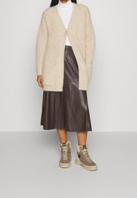 Beige wollen cardigan met open voorkant en zijzakken, gedragen over een witte coltrui en een bruine leren midi-rok, gecombineerd met grijsbruine laarzen.