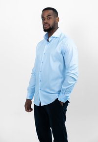 Lichtblauwe blouse met lange mouwen, met een knoopsluiting, gestreept textuur en blauwe knopen. Gecombineerd met donkere broek, casual gedragen.