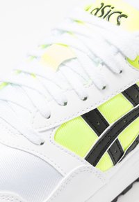 Sneaker bianco realizzato in materiali mesh e sintetici, con accenti giallo neon, dettagli a strisce nere e lacci bianchi con una superficie texturizzata.