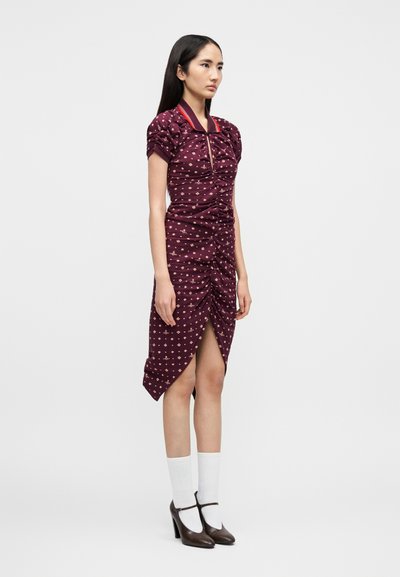 Nuori nainen pukeutuneena purppuranväriseen rypytettyyn mekkoon, jossa on punareunainen kaulus, valkoiset sukat ja ruskeat Mary Jane -korkokengät, seisoo yksinkertaisen taustan edessä.