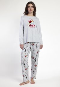 Mujer vestida con un pijama gris de Alicia en el País de las Maravillas con diseños de un conejo blanco y cartas de juego, de pie descalza contra un fondo liso.