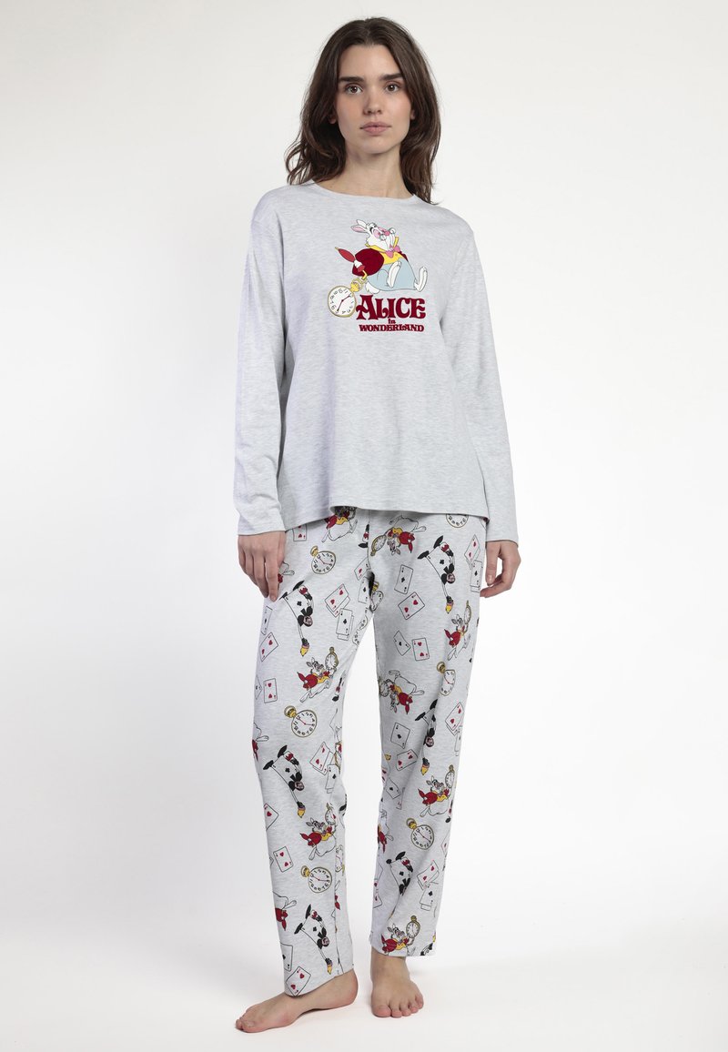 Mujer vestida con un pijama gris de Alicia en el País de las Maravillas con diseños de un conejo blanco y cartas de juego, de pie descalza contra un fondo liso.