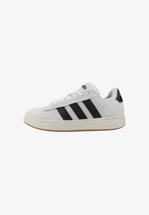 Zapatilla blanca de Adidas con rayas negras, diseño con cordones y suela beige sobre un fondo blanco, vista lateral.
