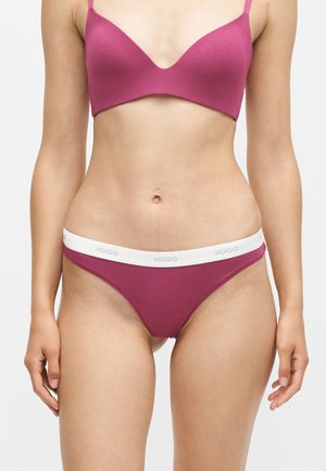 Kvinde iført pink bh og matchende undertøj med hvid linning mærket "HUGO", vist fra nakke til øvre lår mod ensfarvet baggrund.
