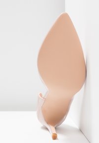 Nude lakované lodičky na vysokém podpatku s špičatou špičkou, tenkým jehlovým podpatkem a hladkým povrchem; minimalistický design na bílém pozadí.