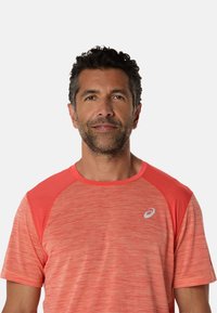 Kortærmet atletisk t-shirt i lys koral med tekstureret stof, raglanærmer og et lille logo på brystet.