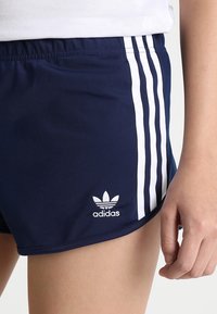 Mörkblå Adidas-shorts med vita tre ränder på sidan och Adidas-logotyp nära fållen, bärs med en vit topp.