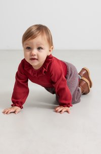 Maglietta lunga a righe rossa con colletto arricciato, abbinata a pantaloni grigi a righe. Bambino che gattona su una superficie liscia di colore chiaro.