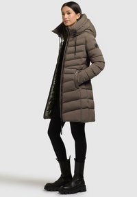 Giacca puffer marrone scuro con cappuccio, caratterizzata da un design trapuntato, chiusura frontale con zip e due tasche laterali. Abbinata a leggins neri e stivali.