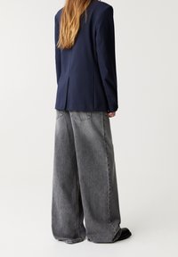 Blazer bleu marine ajusté avec une fente arrière unique, associé à un jean denim gris à jambes larges avec poches arrière et texture délavée.