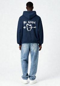 Námornícka mikina s veľkým bielym grafickým nápisom "Mi Amor" a šípkou na zadnej strane; skombinovaná so svetlomodrými voľnými džínsy a bielymi teniskami.