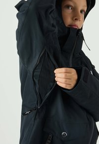 Black waterproof jacket with a zippered opening under the arm, textured fabric, a high collar, and a snap button closure at the hem.