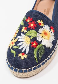 Espadrille bleu marine avec des fleurs brodées rouges, jaunes et blanches ainsi que des feuilles vertes, dotée d'une semelle en jute tressé.