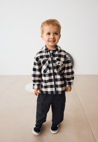 Schwarz-weiß karierte Flanelljacke mit Frontreißverschluss, mit Brusttasche und Pandadetail, kombiniert mit schwarzen Cargohosen und schwarzen Schuhen.