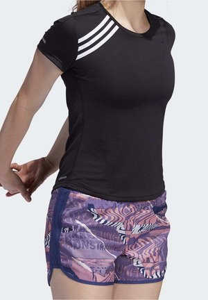 Femme portant un t-shirt noir à manches courtes avec des rayures blanches et un short de sport violet coloré, étirant son bras derrière son dos.
