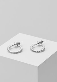 Pendientes de aro de plata con un diseño circular, adornados con pequeñas piedras transparentes a lo largo de un borde. Cuentan con un cierre de tipo stud.