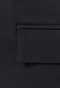 Tissu noir à texture lisse, présentant un détail de poche cousue et des lignes épurées, adapté aux tenues formelles ou professionnelles.