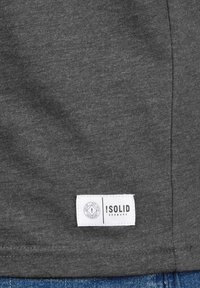 Dunkelgraues Baumwoll-T-Shirt mit einem weißen Markenetikett und schwarzem Text sowie Logo auf der unteren linken Seite. Glatte Textur, lässiges Design.
