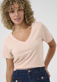 Ljusrosa t-shirt med v-ringning och korta ärmar, tillverkad av mjukt tyg, i kombination med mörkblå högmidjade denimshorts med metallknappdetaljer.